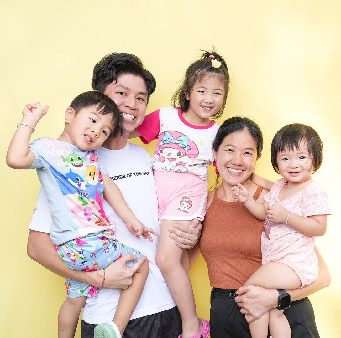 A cheerful family wearing matching pastel clothing poses together against a yellow wall background.