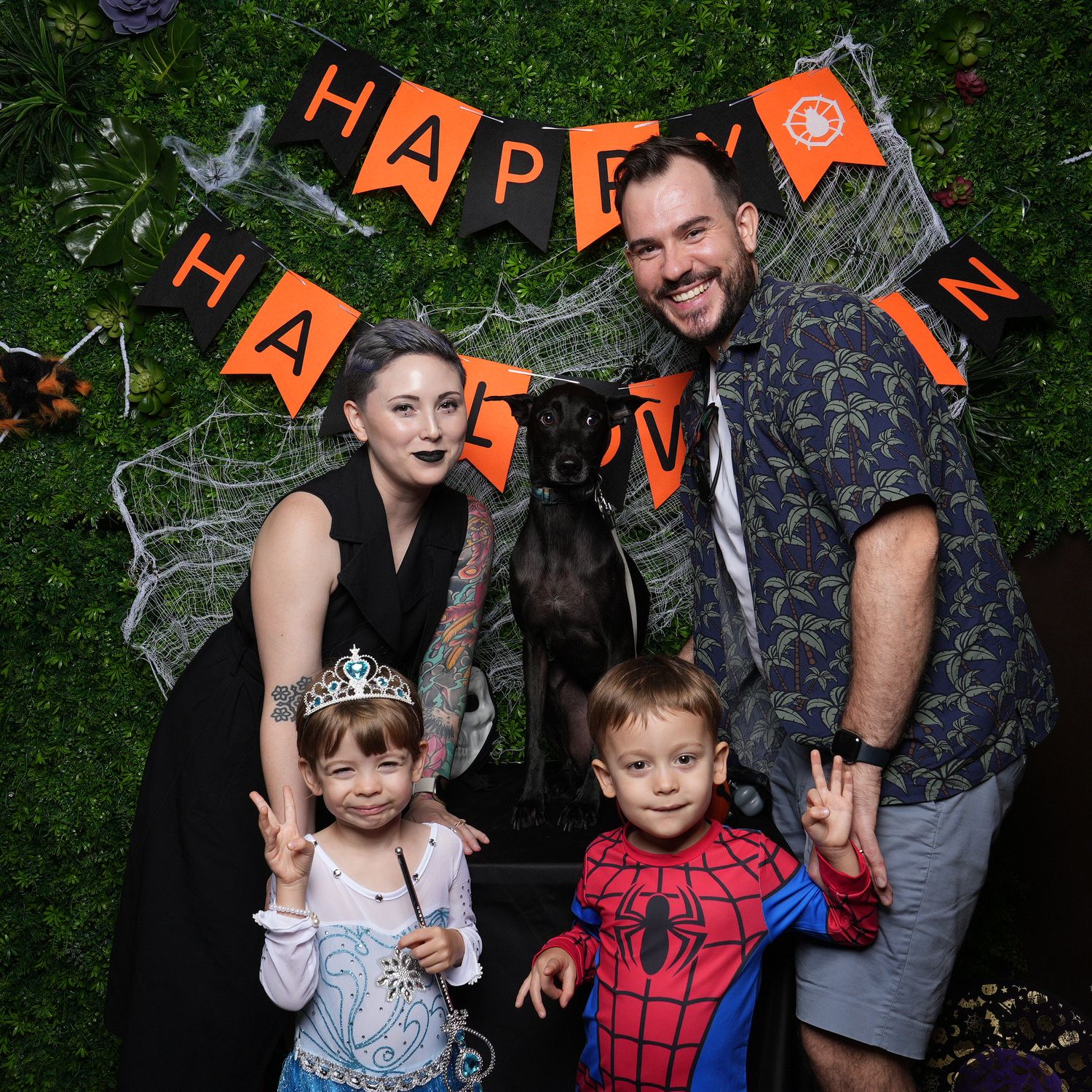 A family dressed in Halloween costumes with princess and Spider-Man outfits pose in front of a spooky decorated green wall.