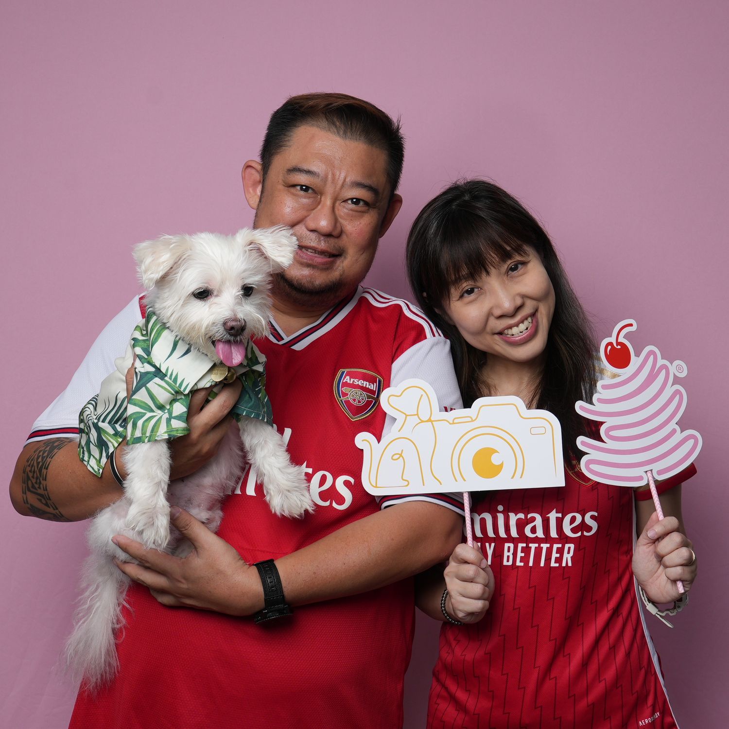 Two Arsenal fans in red jerseys pose with a small white dog in clothing and hold up cartoon cutouts of ice cream.