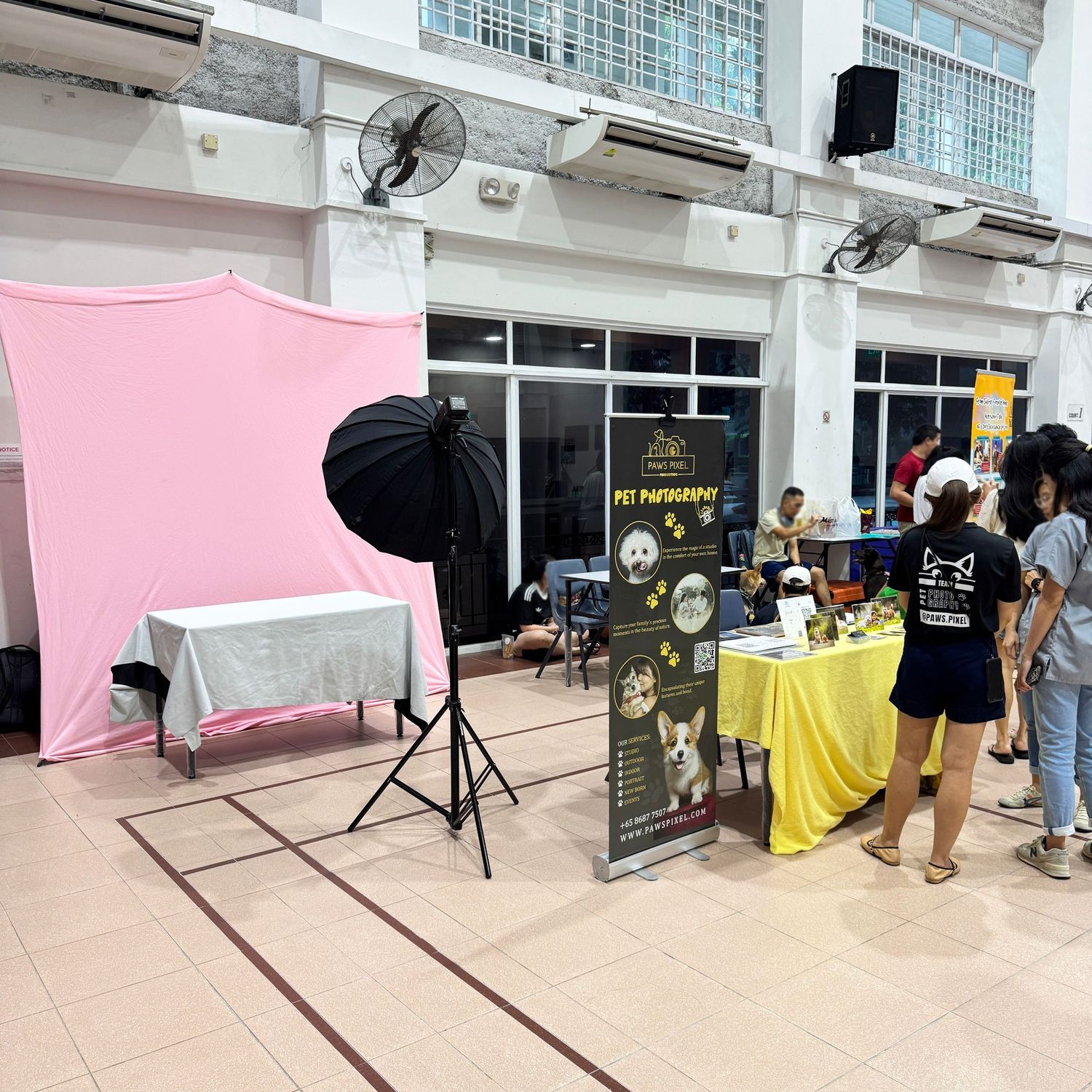 A photography setup with pink backdrop and lighting equipment next to vendor displays at an outdoor market or event area.