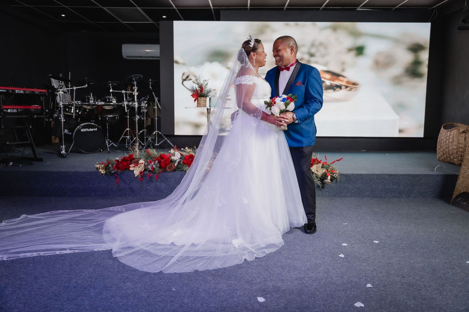 Newlywed couple shares an intimate moment on their wedding day in an indoor venue with scattered red roses on the floor.