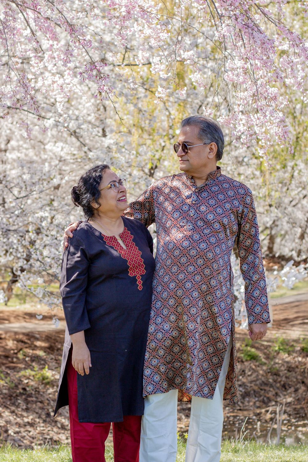 South Asian couple in traditional attire smiling together outdoors with blooming cherry blossom trees in background.