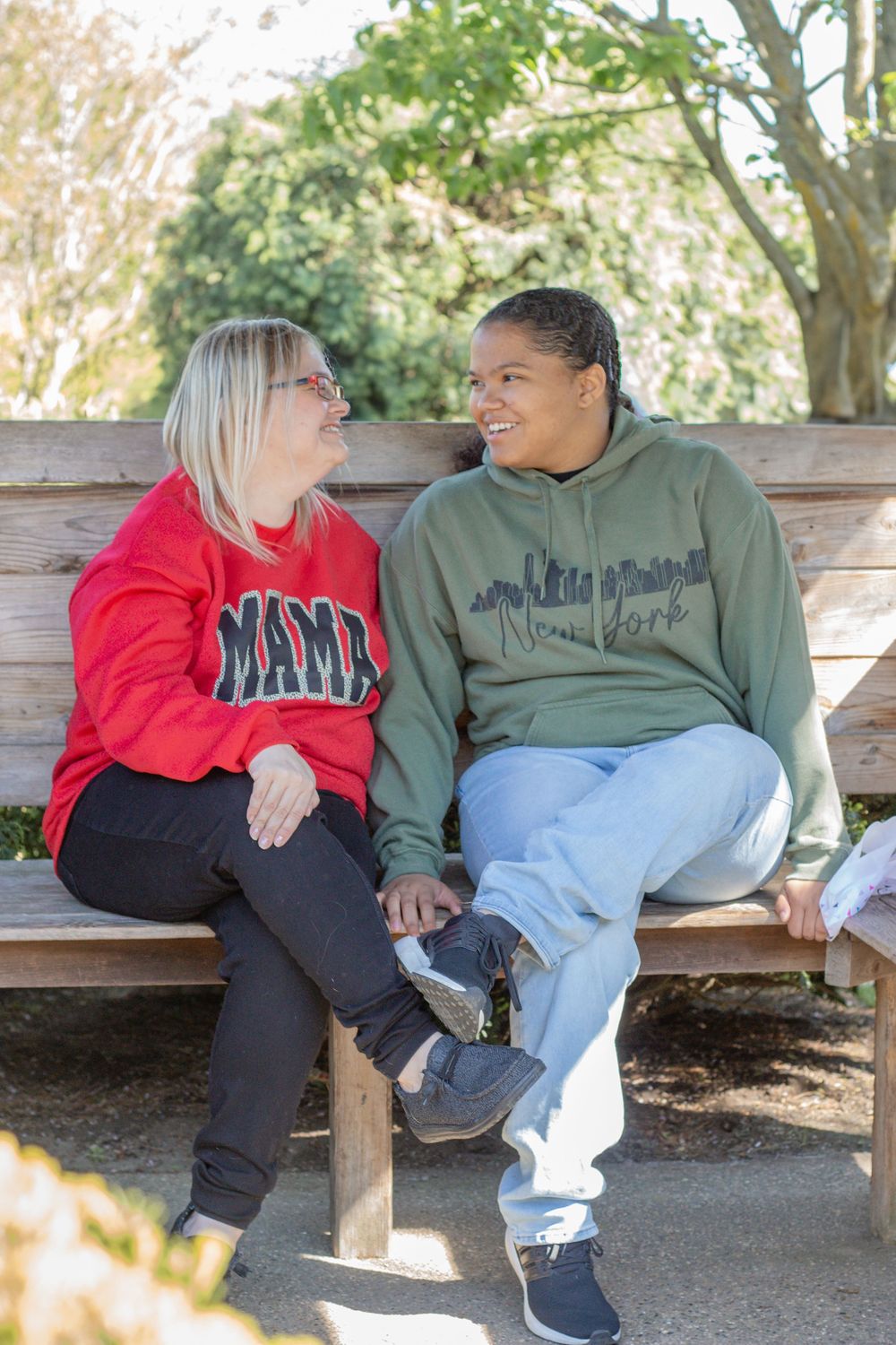 Two women sitting on a wooden bench outdoors, smiling at each other, one in a red MAMA sweatshirt, one in a green New York hoodie.
