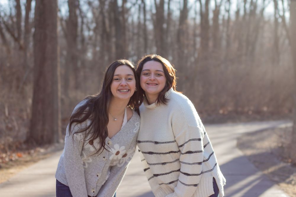 Two young women smiling together on a tree-lined road in autumn, wearing cozy sweaters outdoors.