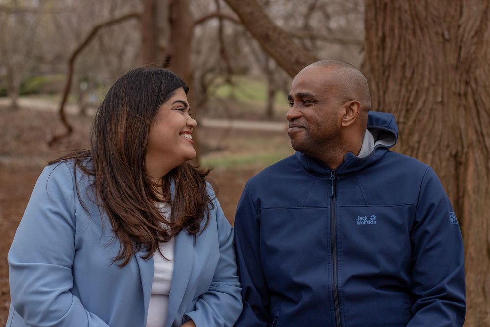 A couple smiles at each other while sitting outdoors in a park surrounded by trees.