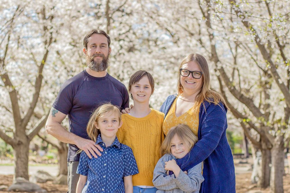 Family of five smiling together outdoors among blooming white cherry blossom trees in spring.