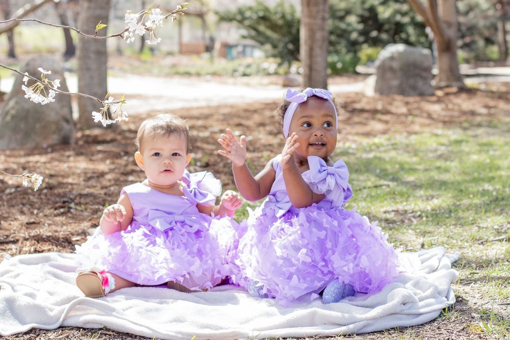Two baby girls in matching purple tutu dresses sit on a white blanket outdoors in a park setting.