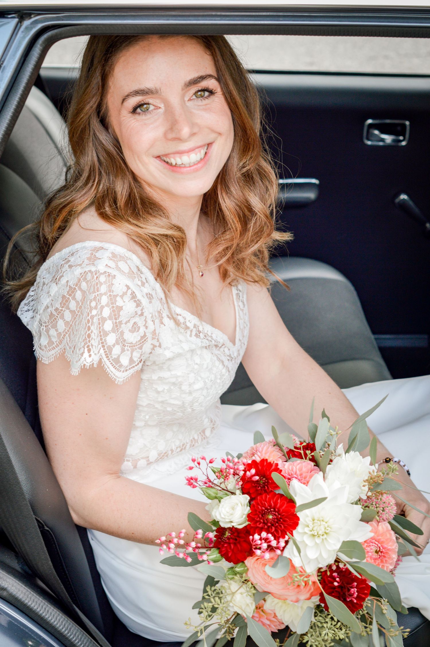 Schöne Braut im Spitzenkleid hält leuchtend roten und weißen Blumenstrauß, während sie im Auto sitzt.