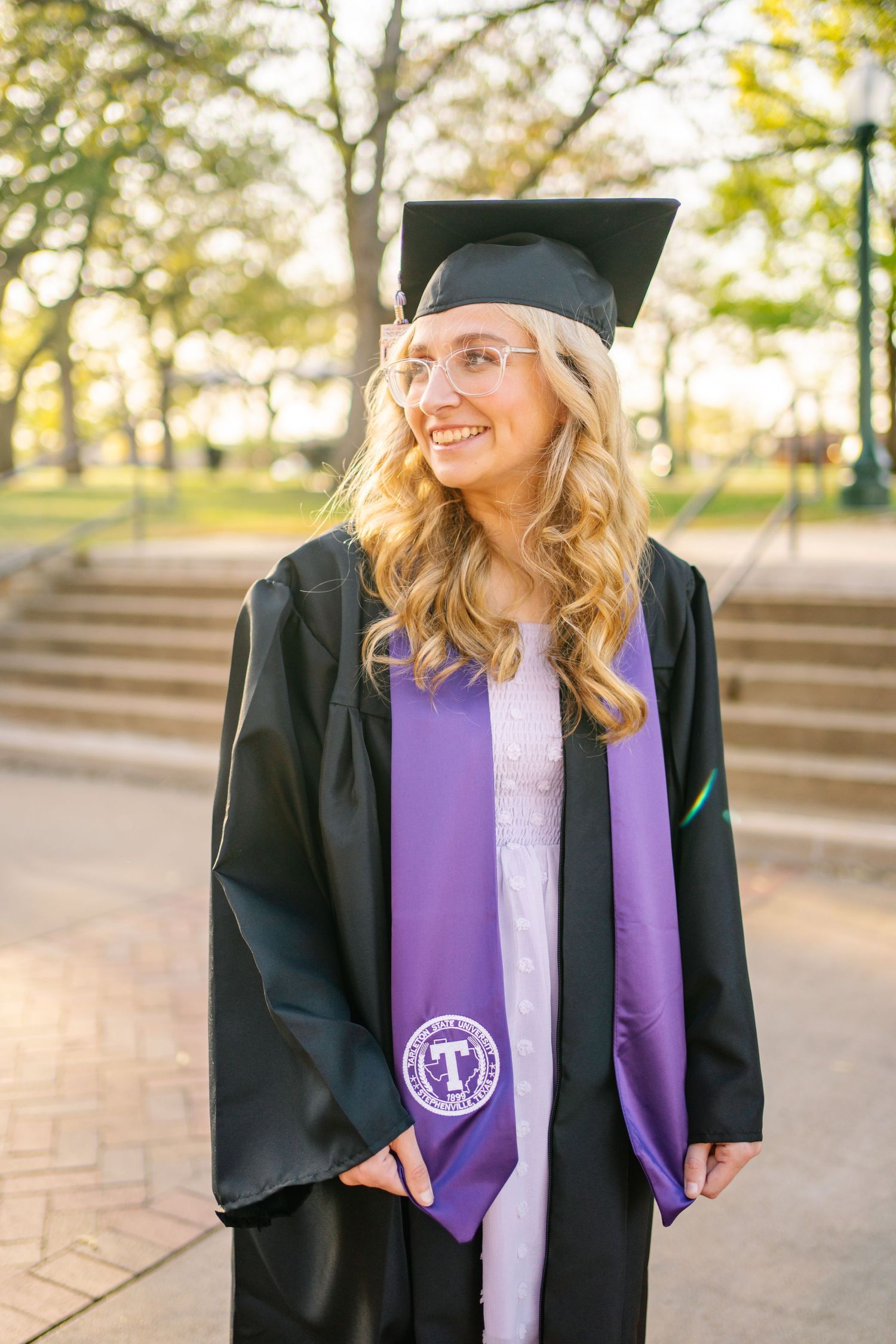 Graduate wearing black cap and gown with purple stole smiles during outdoor graduation photoshoot.