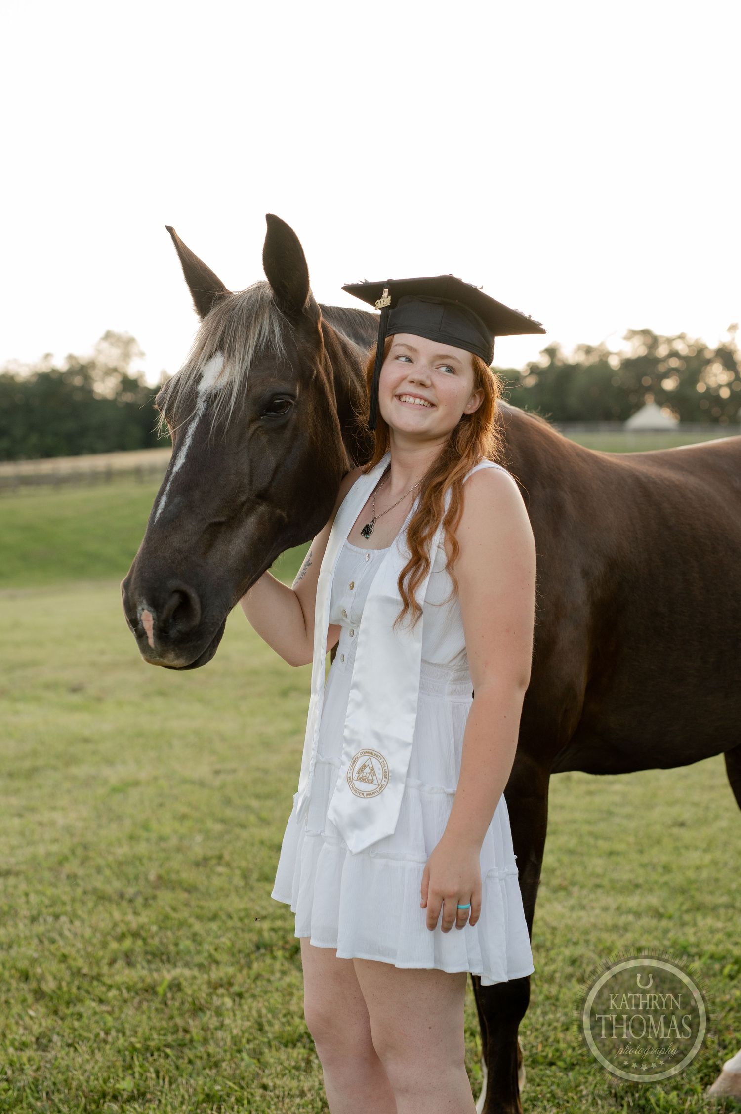 Graduate poses with dark horse in field while wearing white dress and graduation stole.
