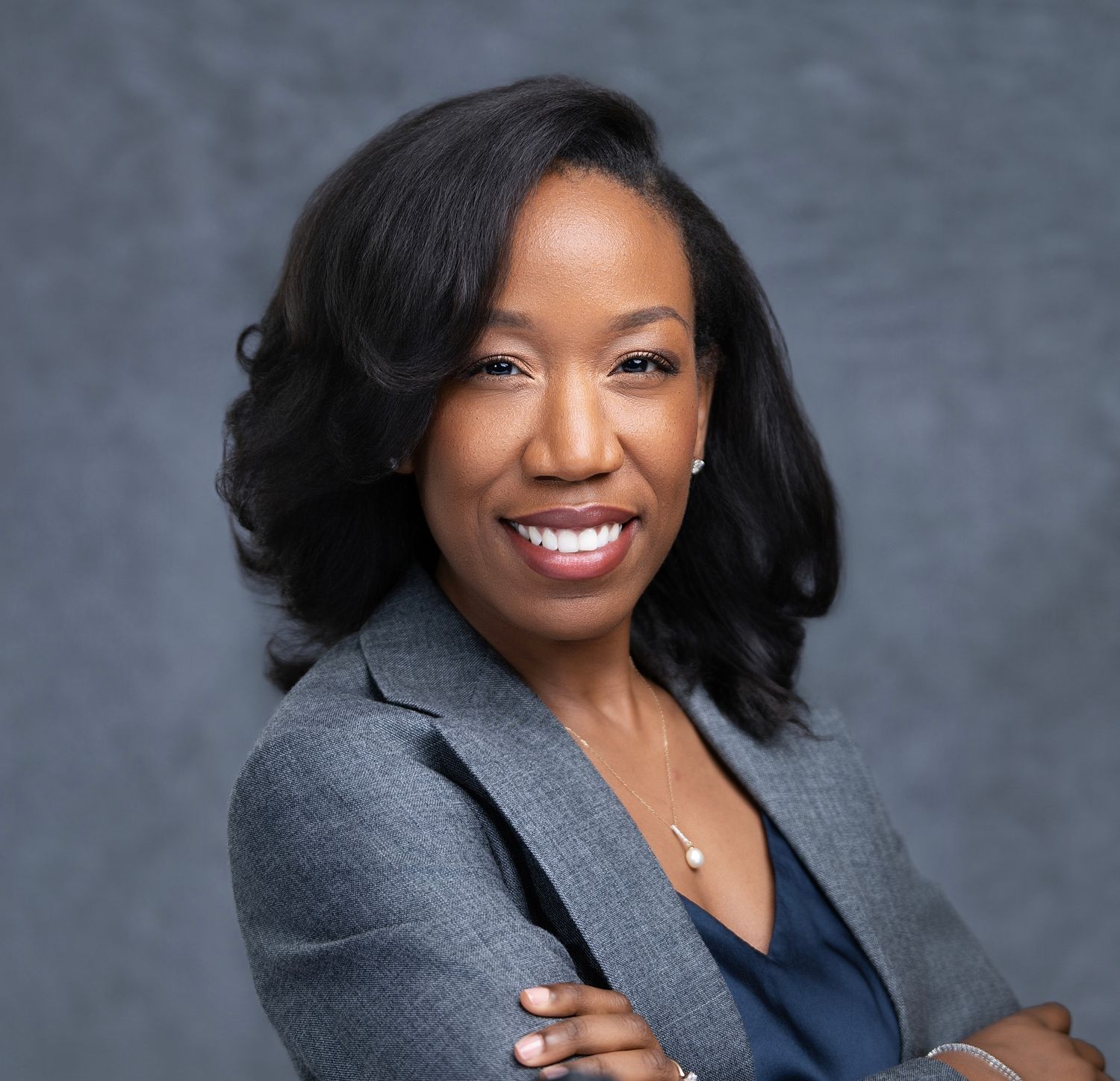 Headshot Professional Black woman in gray blazer smiling with arms crossed against gray background.