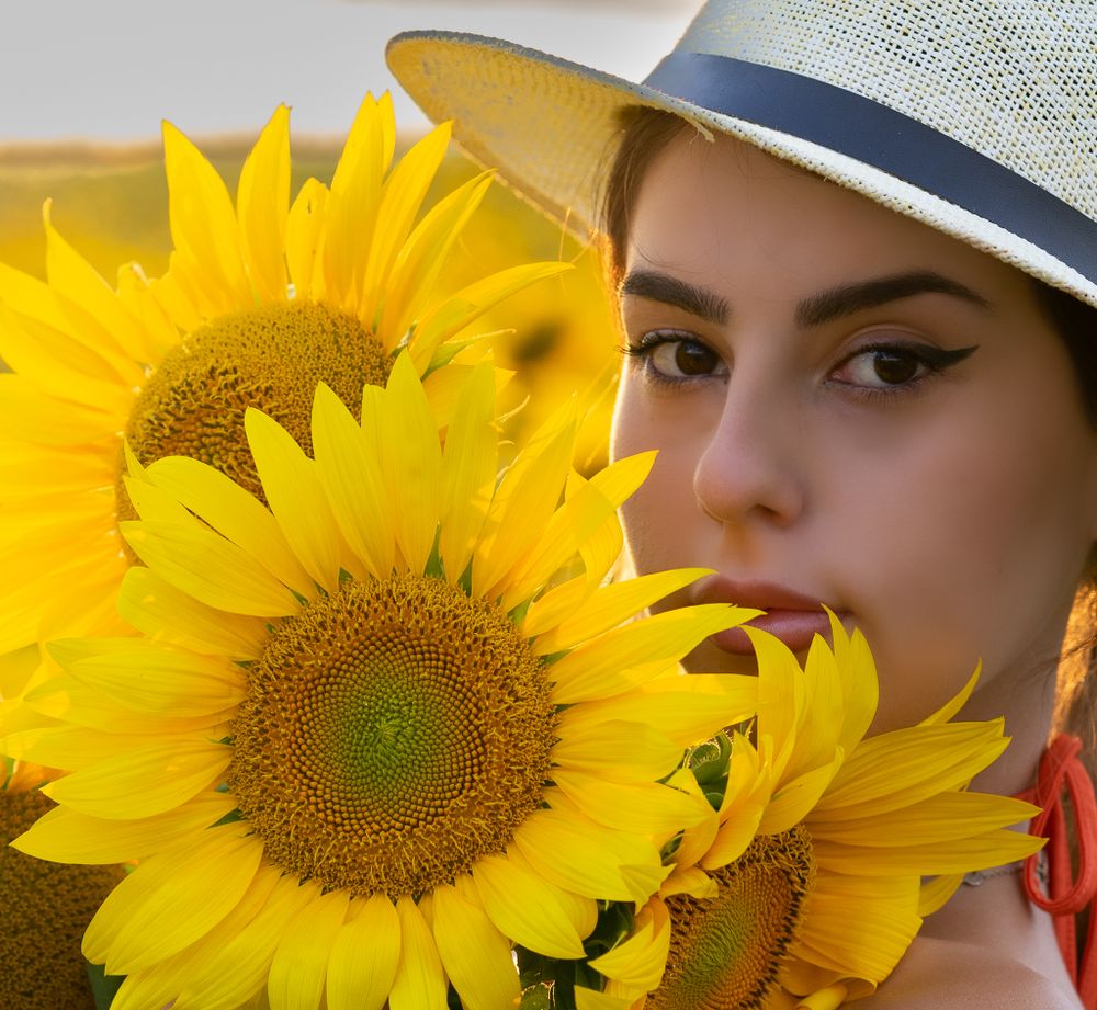 Beautyfotografie - a girl with bright yellow sunflowers contrast with summer hat in outdoor portrait.