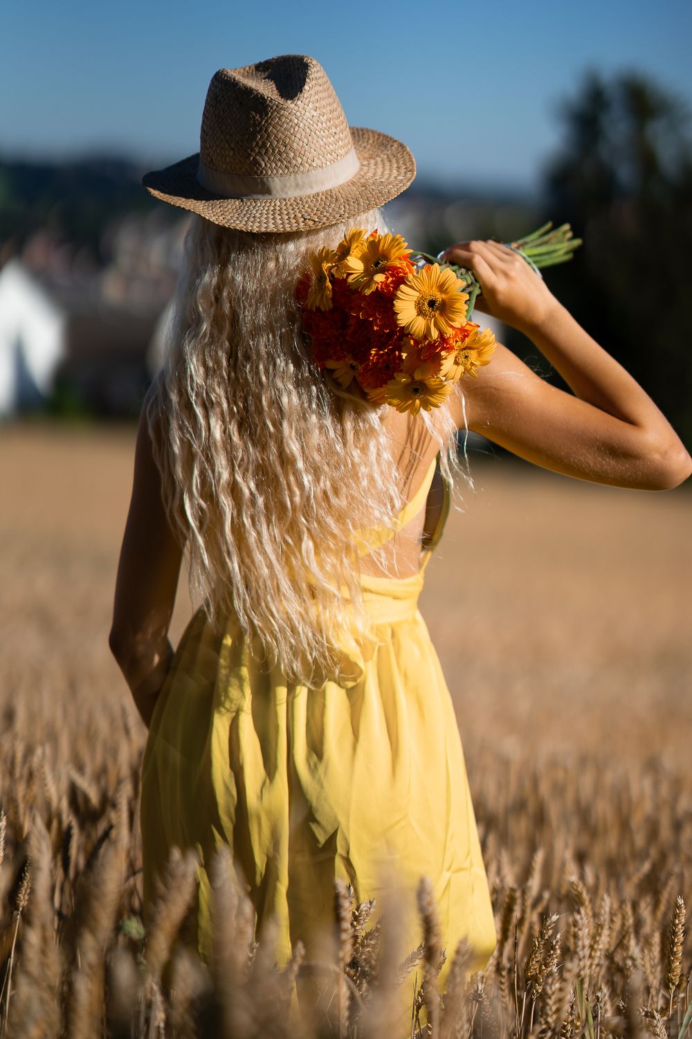 Beauty Portrait of a woman in yellow dress and straw hat holding flowers in wheat field at sunset.