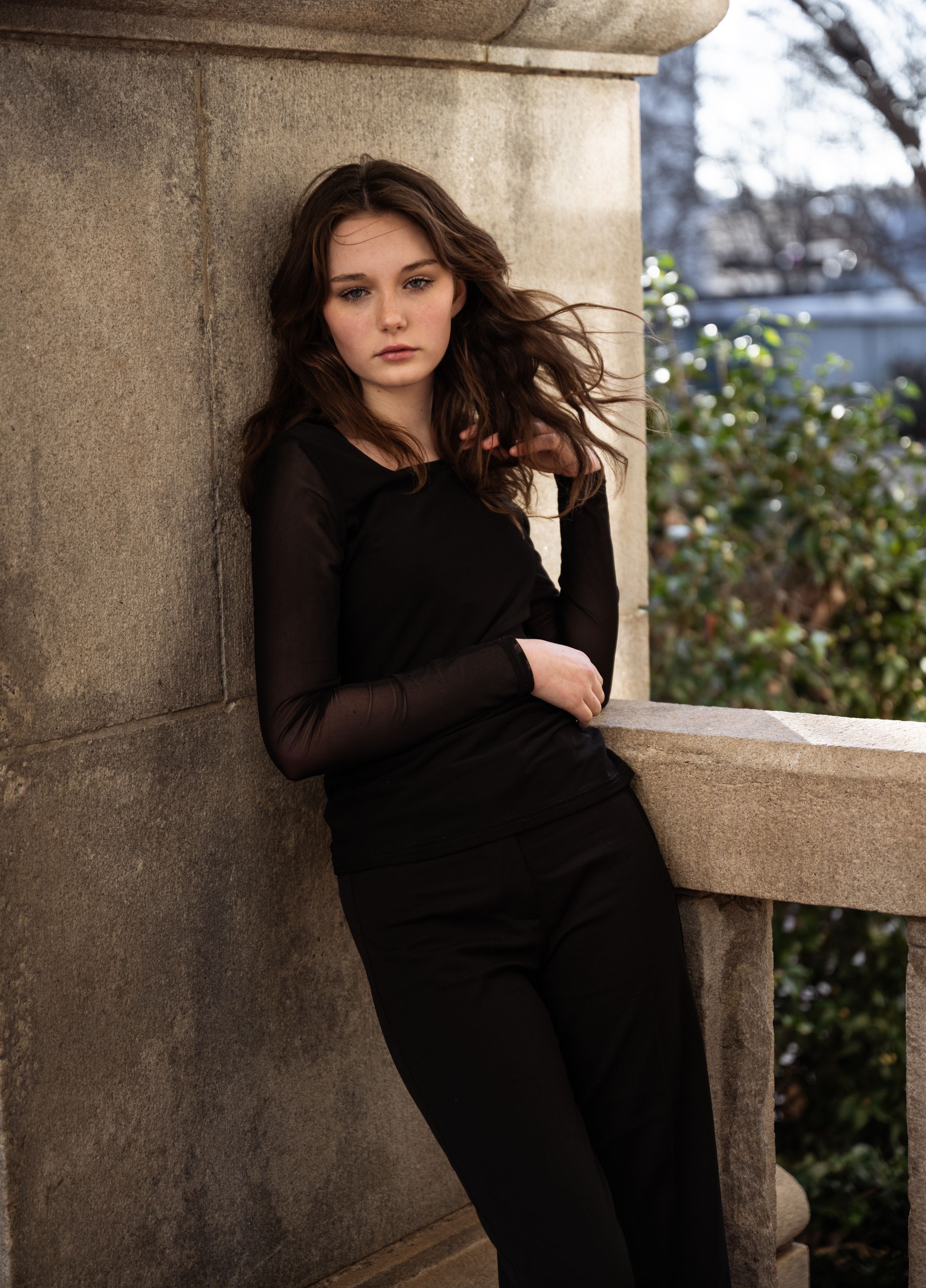 An artistic portrait of a woman in black attire leaning against a stone column outdoors.