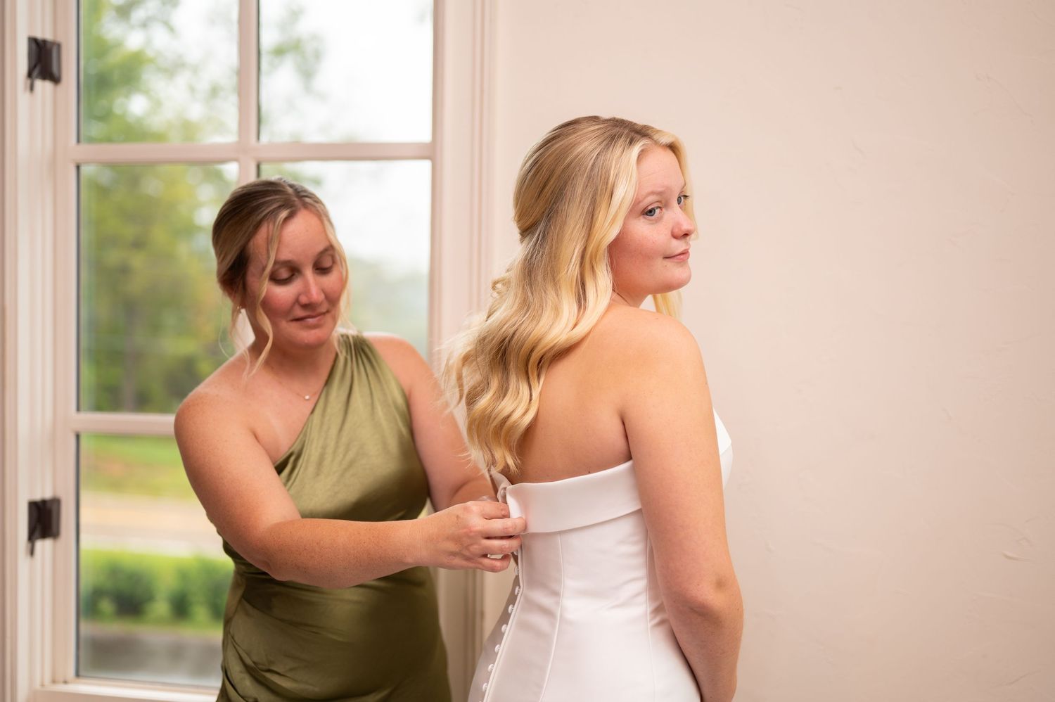 A bridesmaid in an olive green dress helps adjust a white wedding gown in front of a sunlit window.