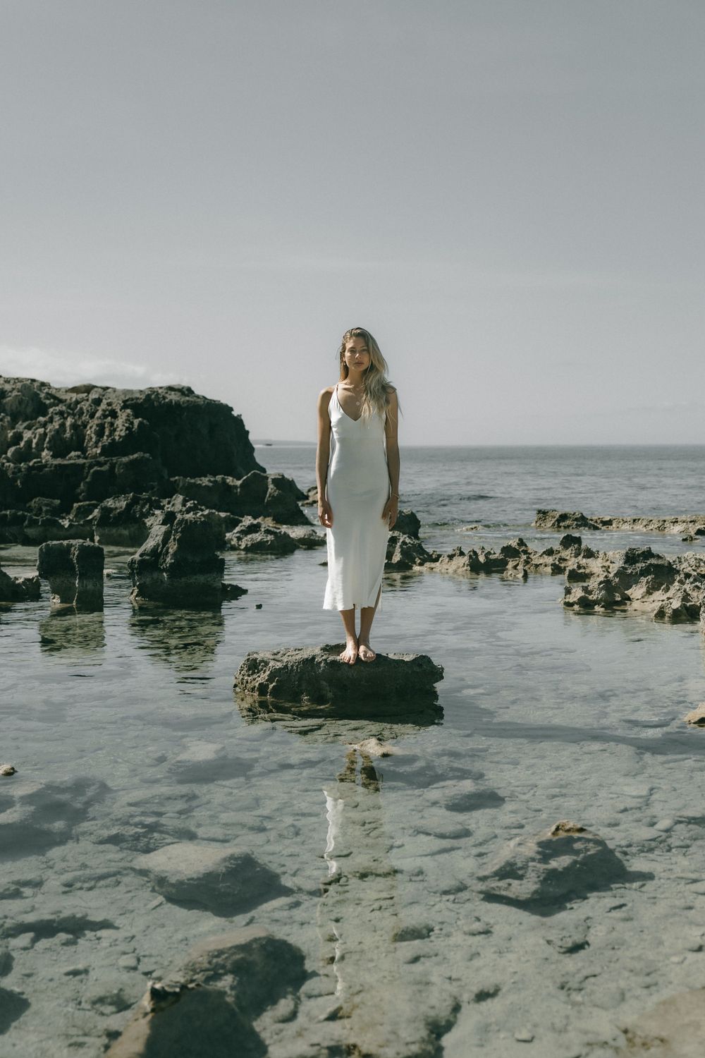 Person in white dress standing on rocky beach landscape with ocean view.