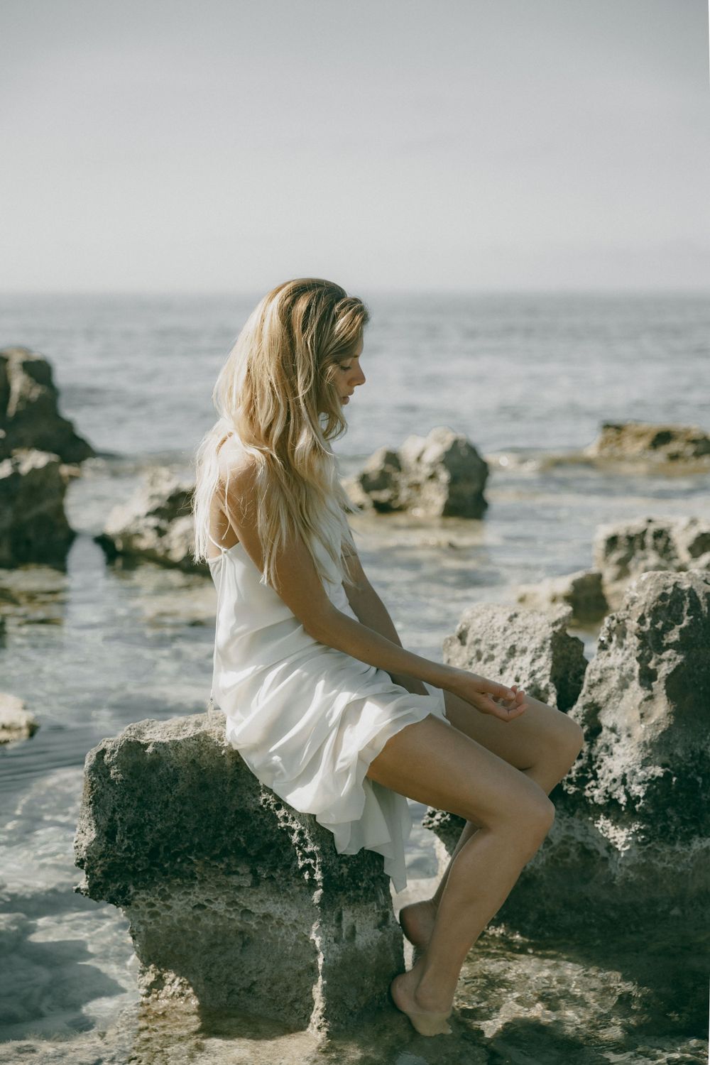 Figure in a white dress sitting on coastal rocks with ocean waves in the background.