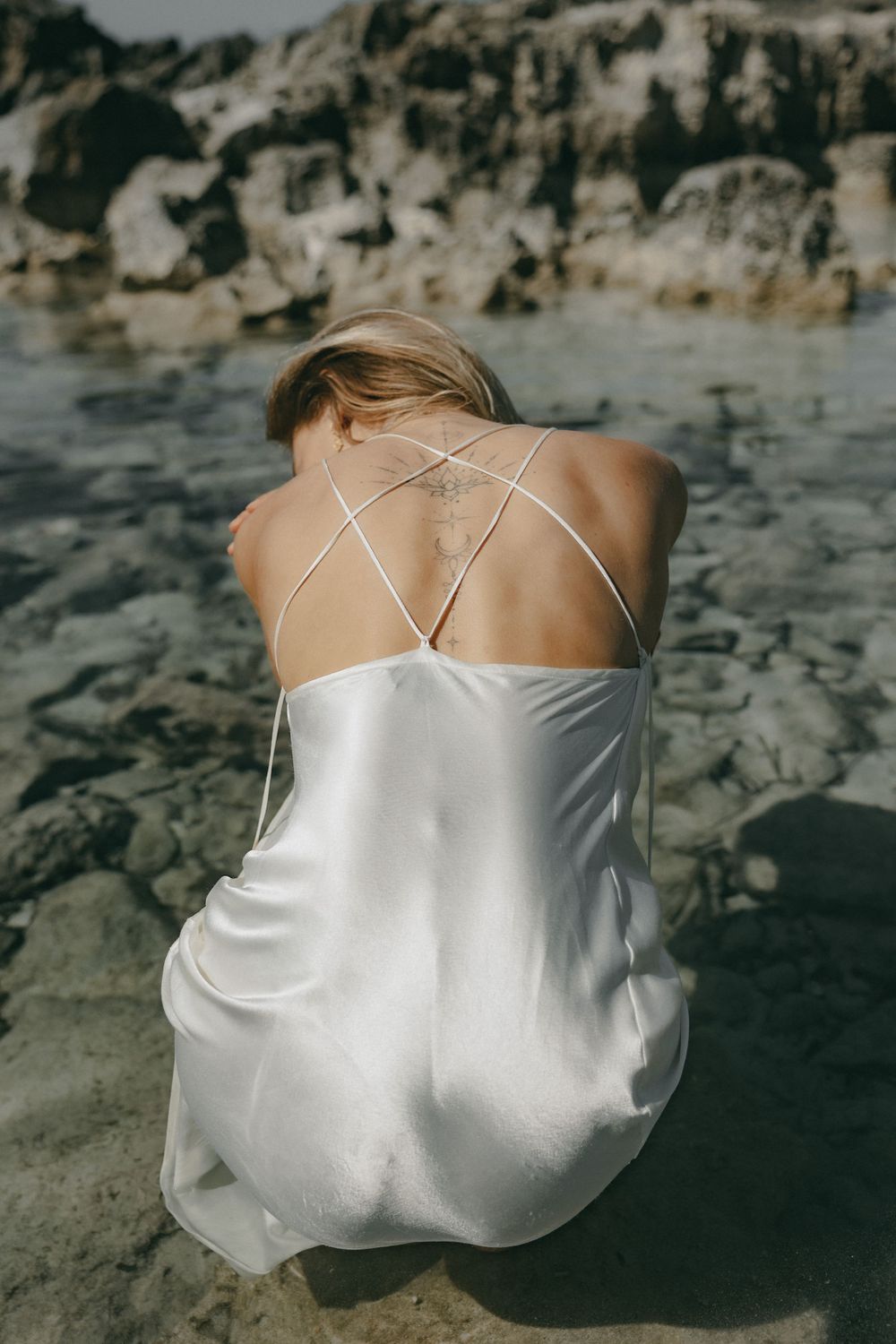 Close-up view of a white silk dress with delicate criss-cross straps against a rocky beach background.
