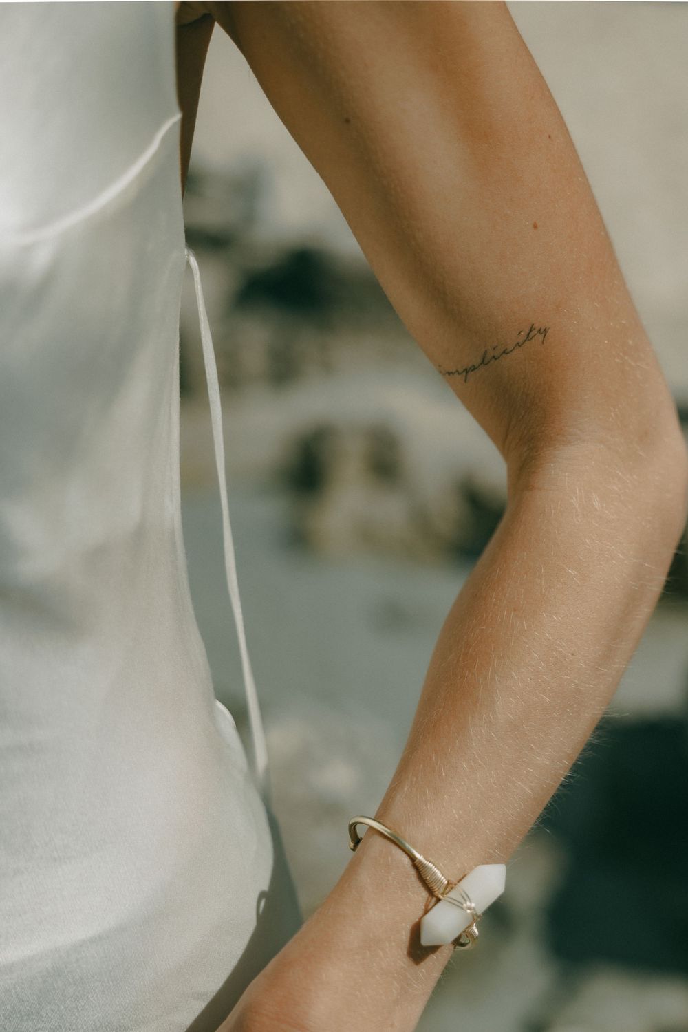 Close-up view of an arm with delicate tattoo and gold bracelet against white fabric.
