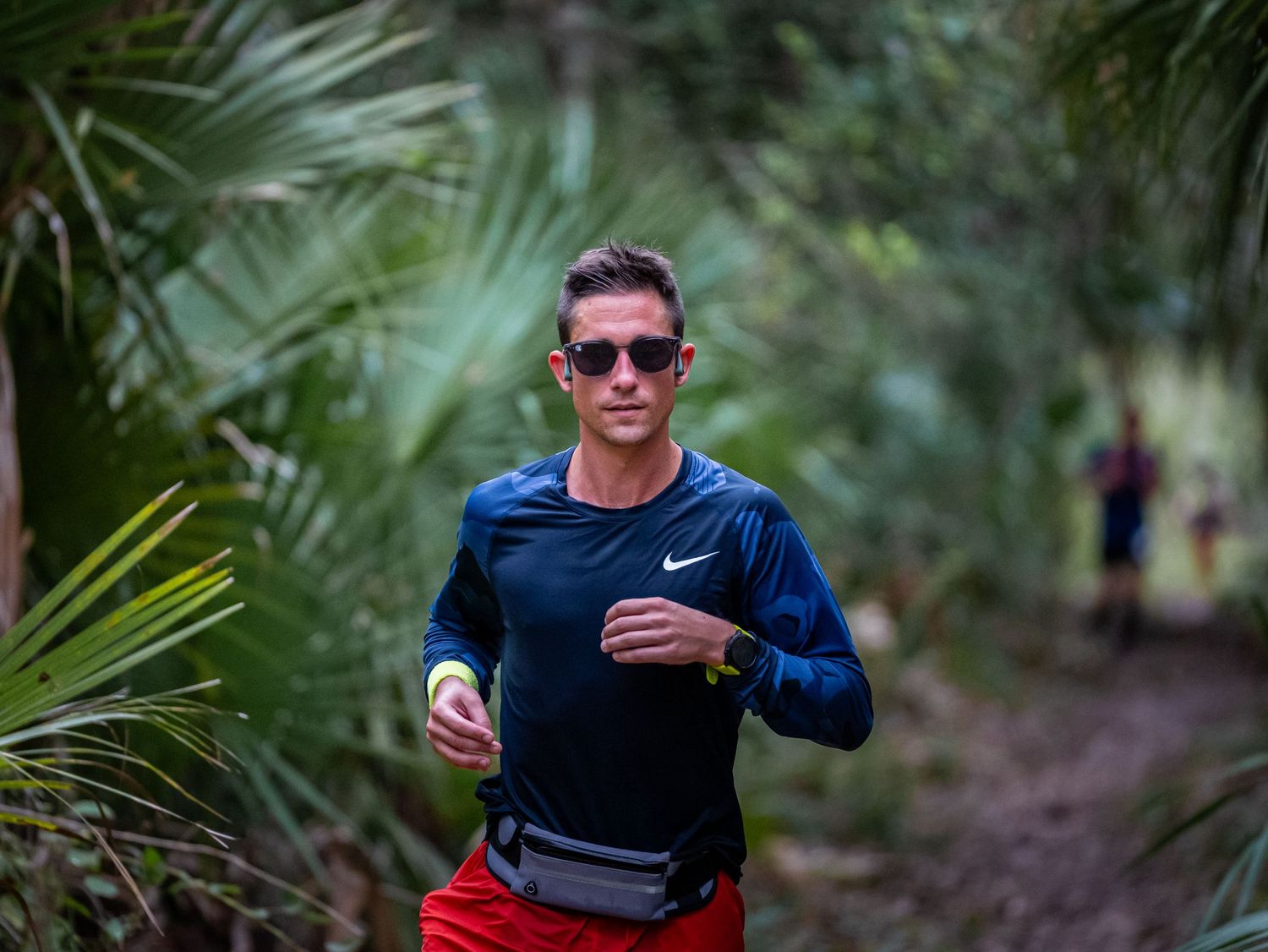 Runner in blue jacket and sunglasses on wooded trail surrounded by palm fronds.