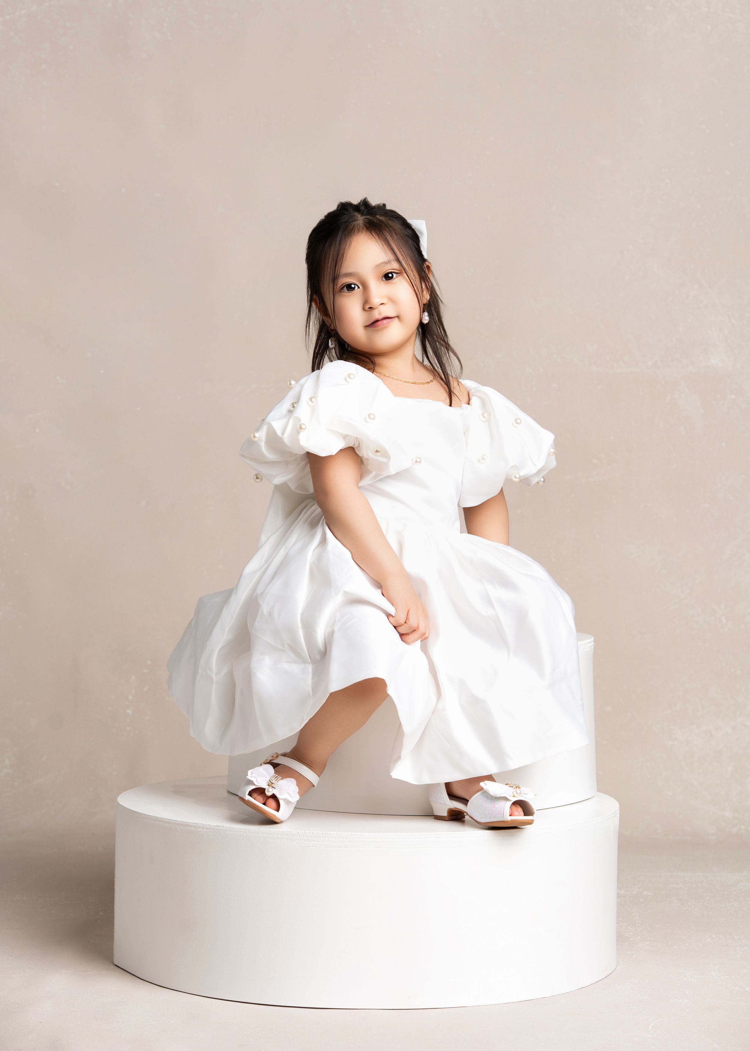 A child in a white puffy dress and sandals poses on a white circular platform against a neutral backdrop.