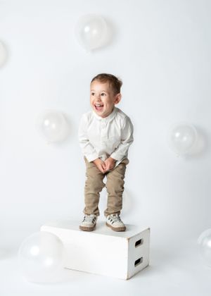 Happy child in white shirt and tan pants stands on white box with transparent balloons floating in minimalist studio birthday photoshoot