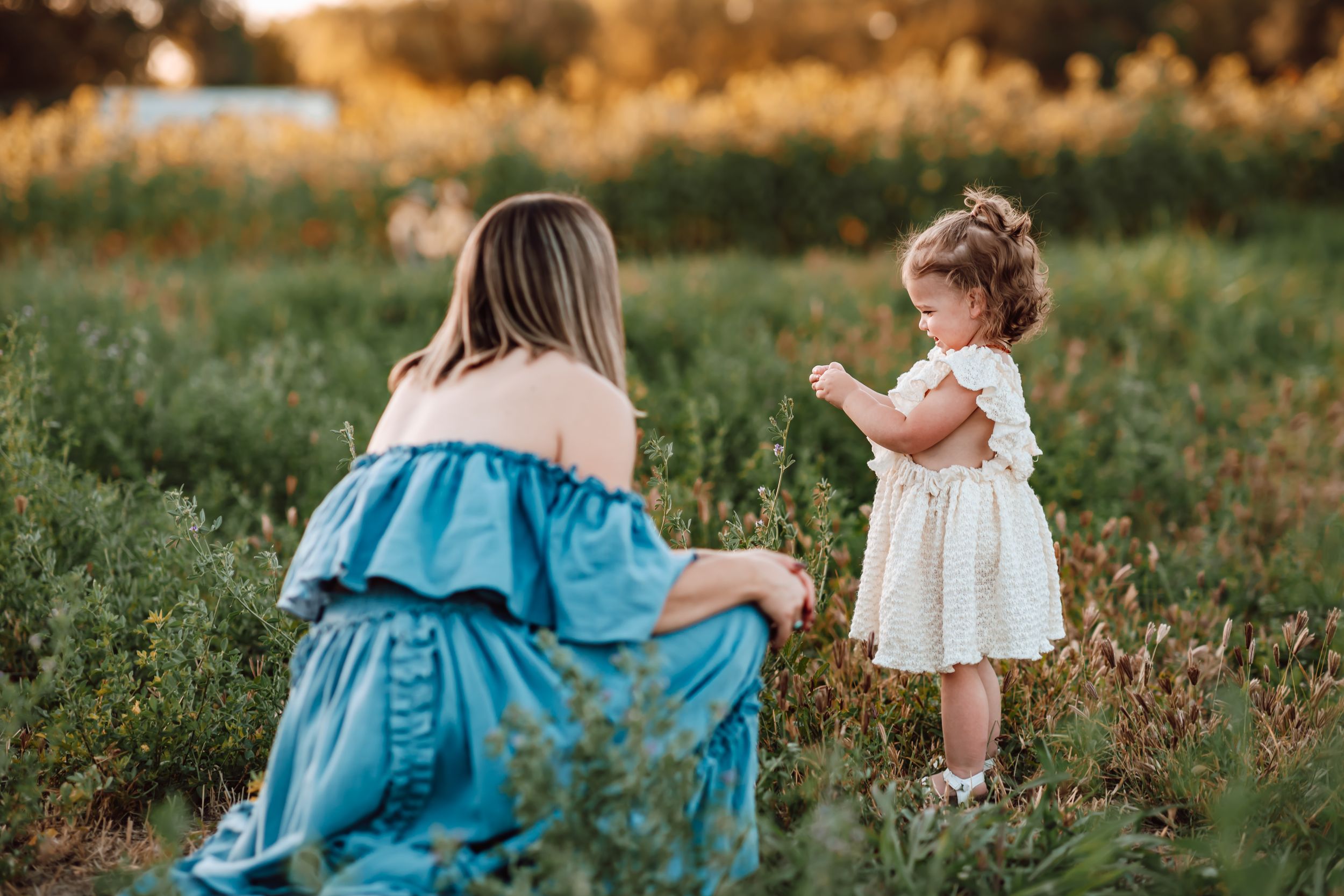 Tender outdoor moment captured at golden hour in a meadow between a woman in blue dress and small child in white.