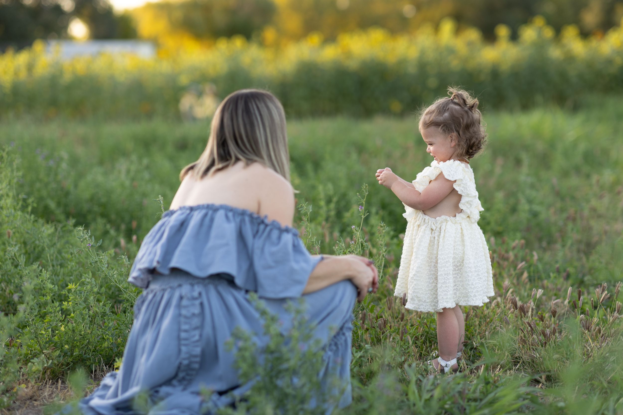 Child in white dress and woman in blue dress share a tender moment together in a sunlit meadow of wildflowers.