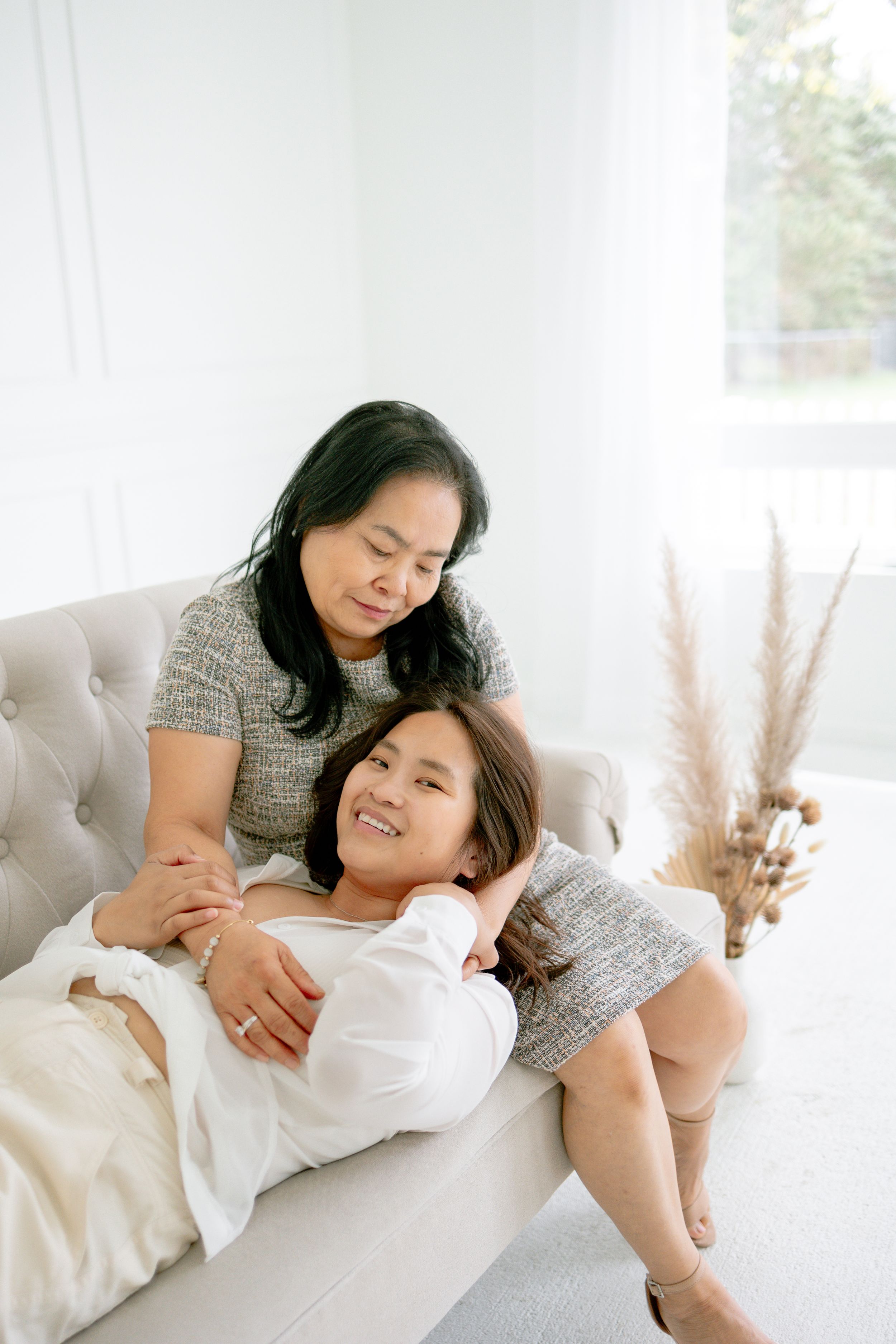 Two people share a tender moment relaxing together on a light gray couch with pampas grass decor in the background.