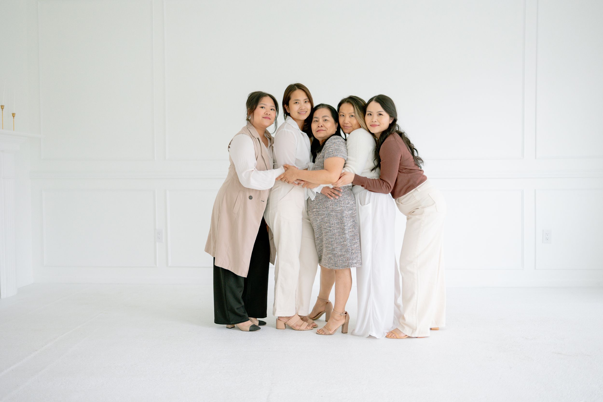 A group of five people wearing neutral-toned outfits embrace while standing against a white backdrop in a studio setting.