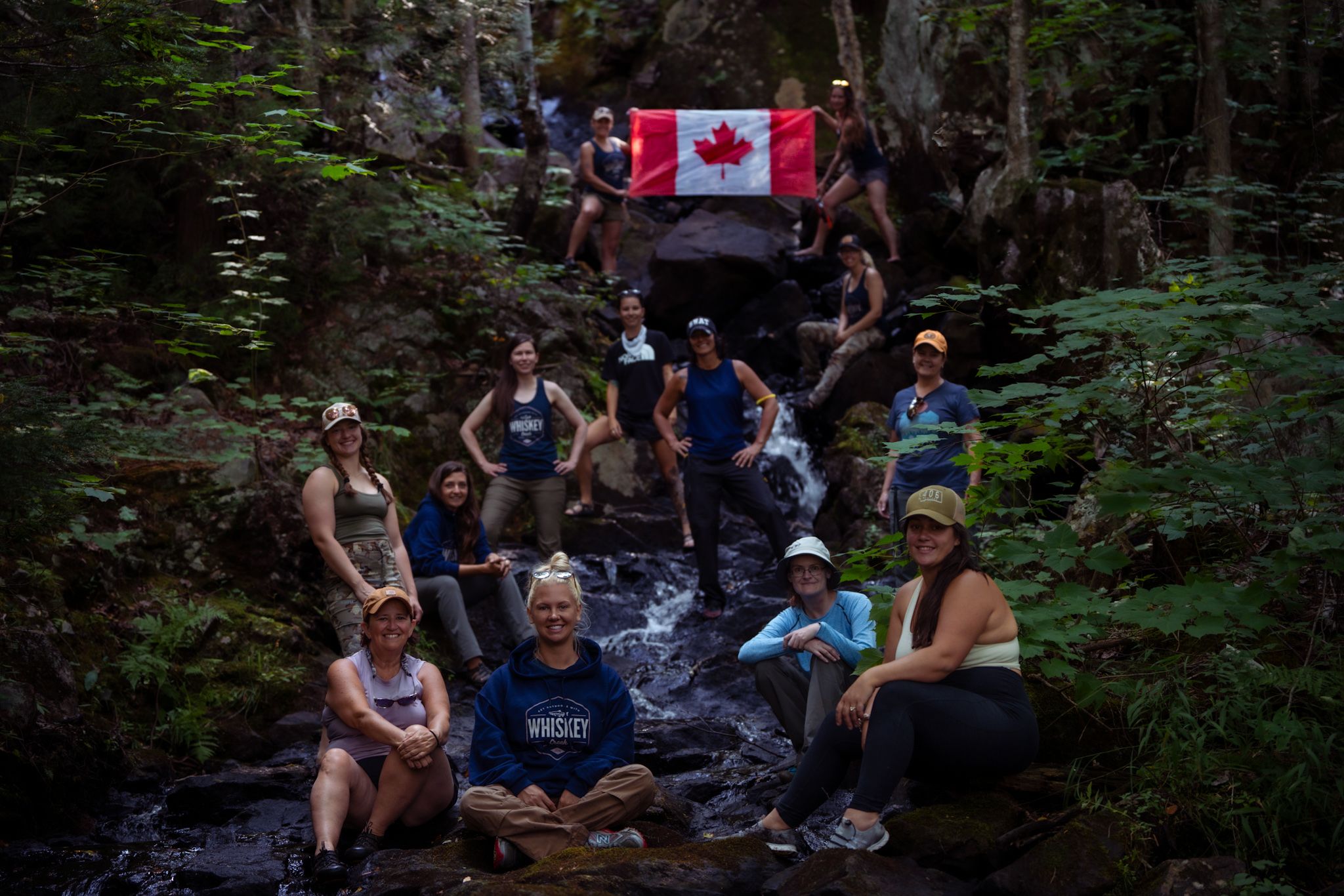A group of hikers rests on rocks by a stream in the woods with a Canadian flag displayed above them.