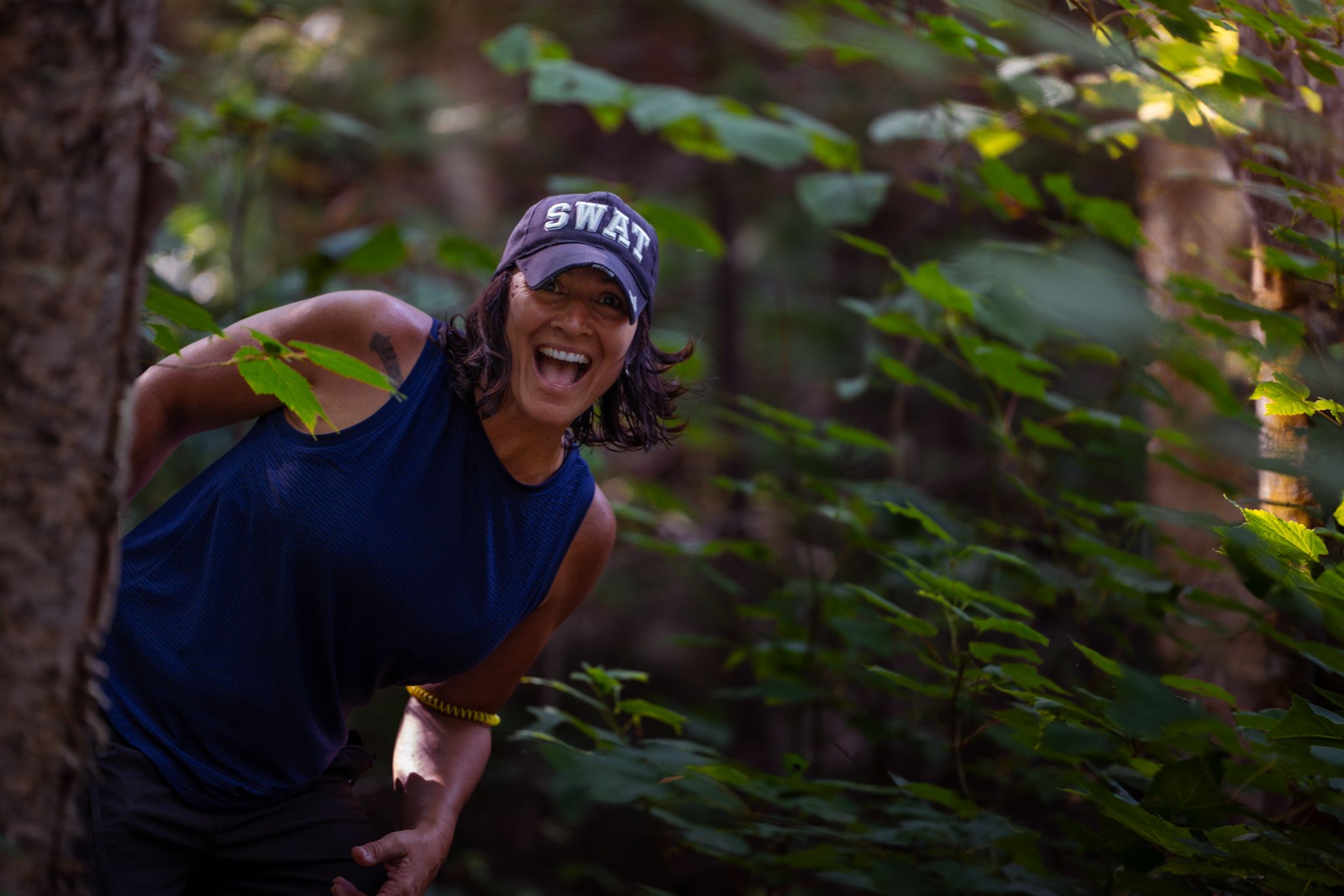 Someone smiling in a SWAT cap and navy shirt in a sunlit forest setting.