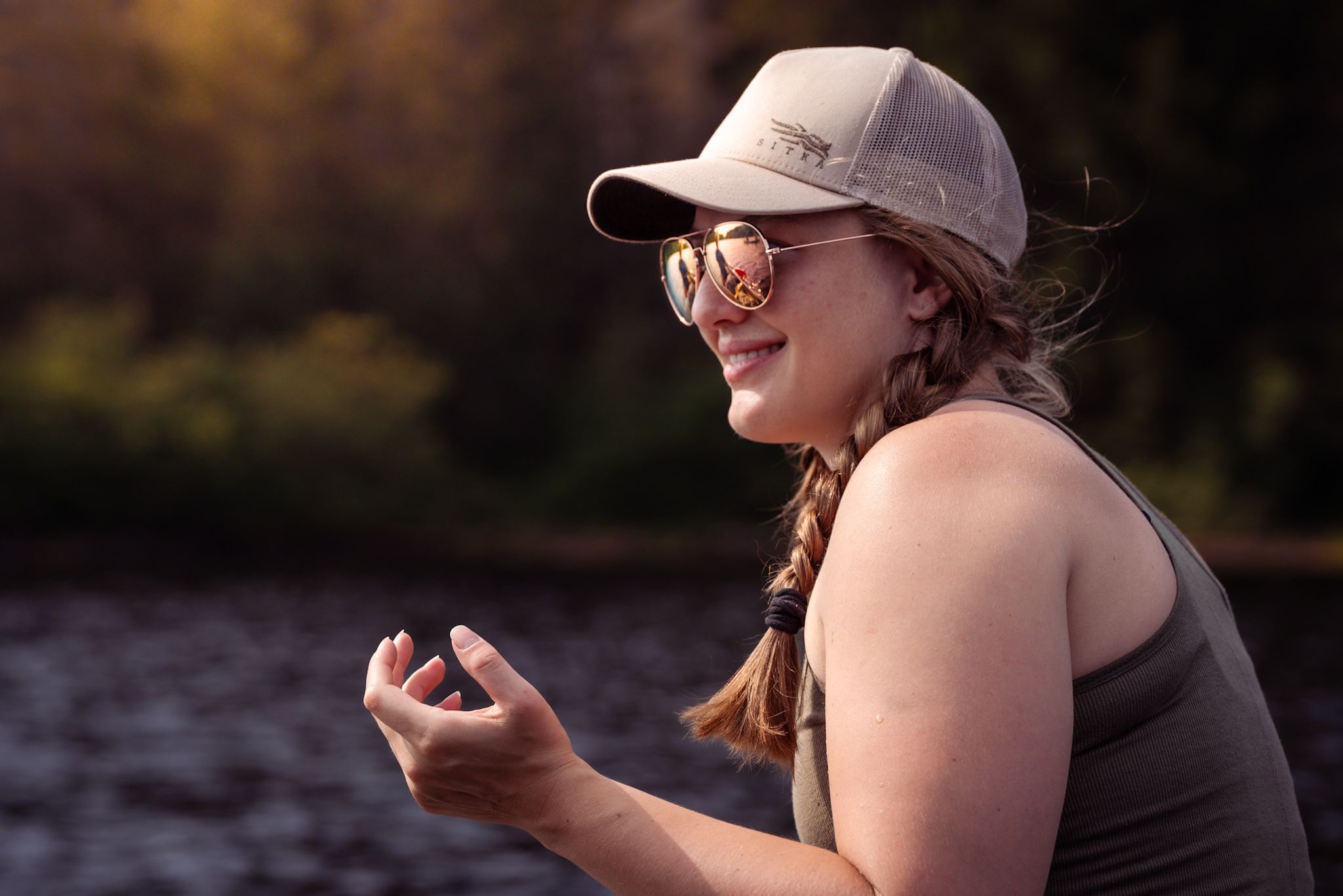 A smiling person in a baseball cap and sunglasses sits by a lake at golden hour.