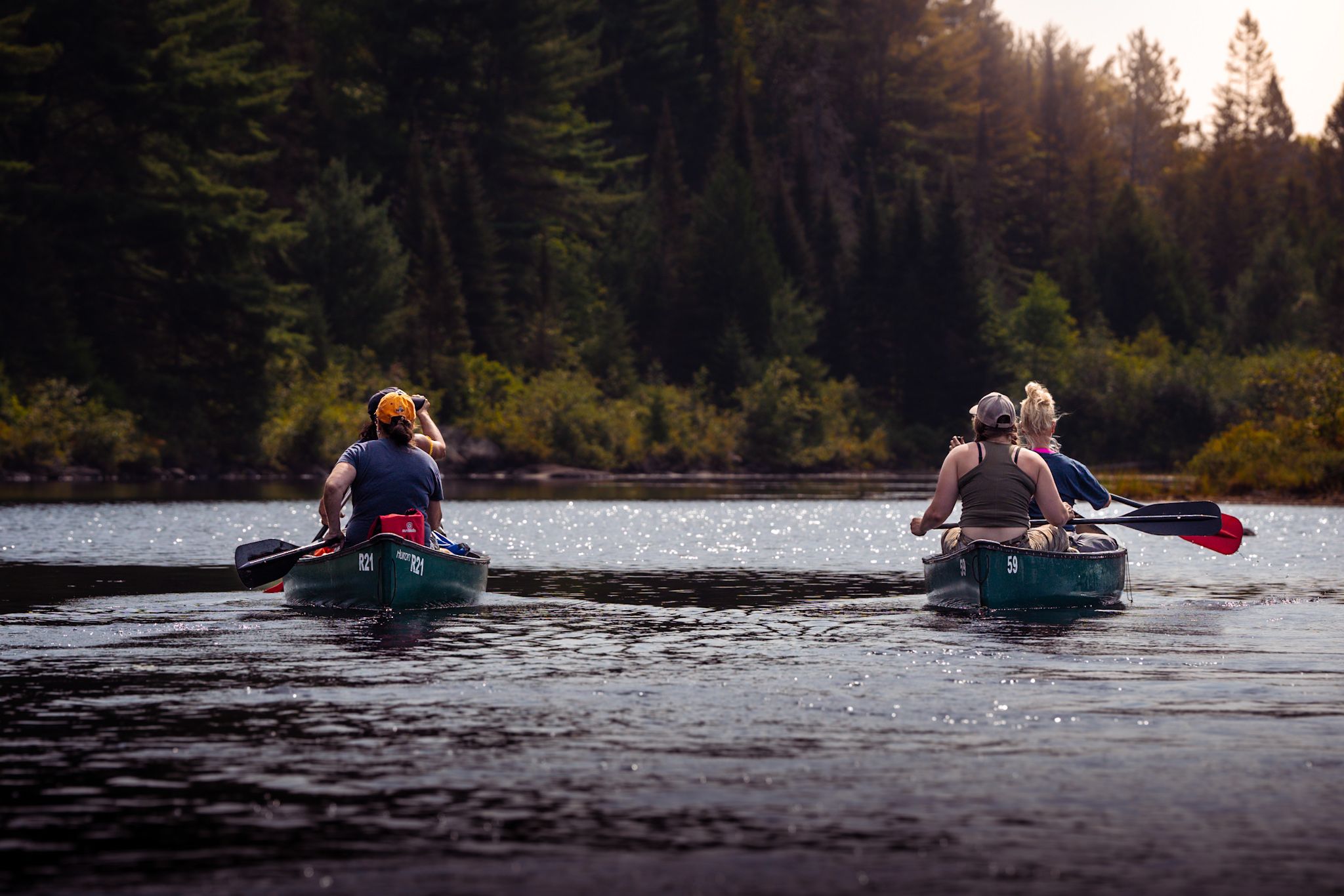 Two kayaks drift along a serene forest lake during golden hour with paddlers visible in silhouette.