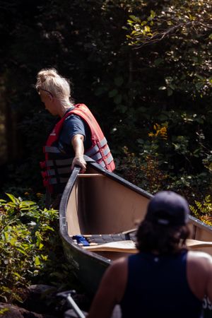 Canoe adventurers in orange life vests prepare their boat for a journey through wooded surroundings on a sunny day.