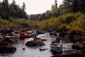 Canoes navigate through rocky rapids in a scenic forest river surrounded by autumn foliage.