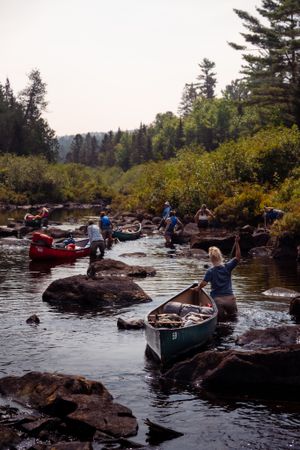 Canoers navigate through rocky waters on a scenic river surrounded by evergreen forest and autumn foliage.