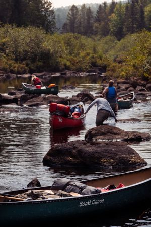 Canoes navigate through rocky waters in a scenic wilderness river surrounded by autumn foliage and evergreen forest.