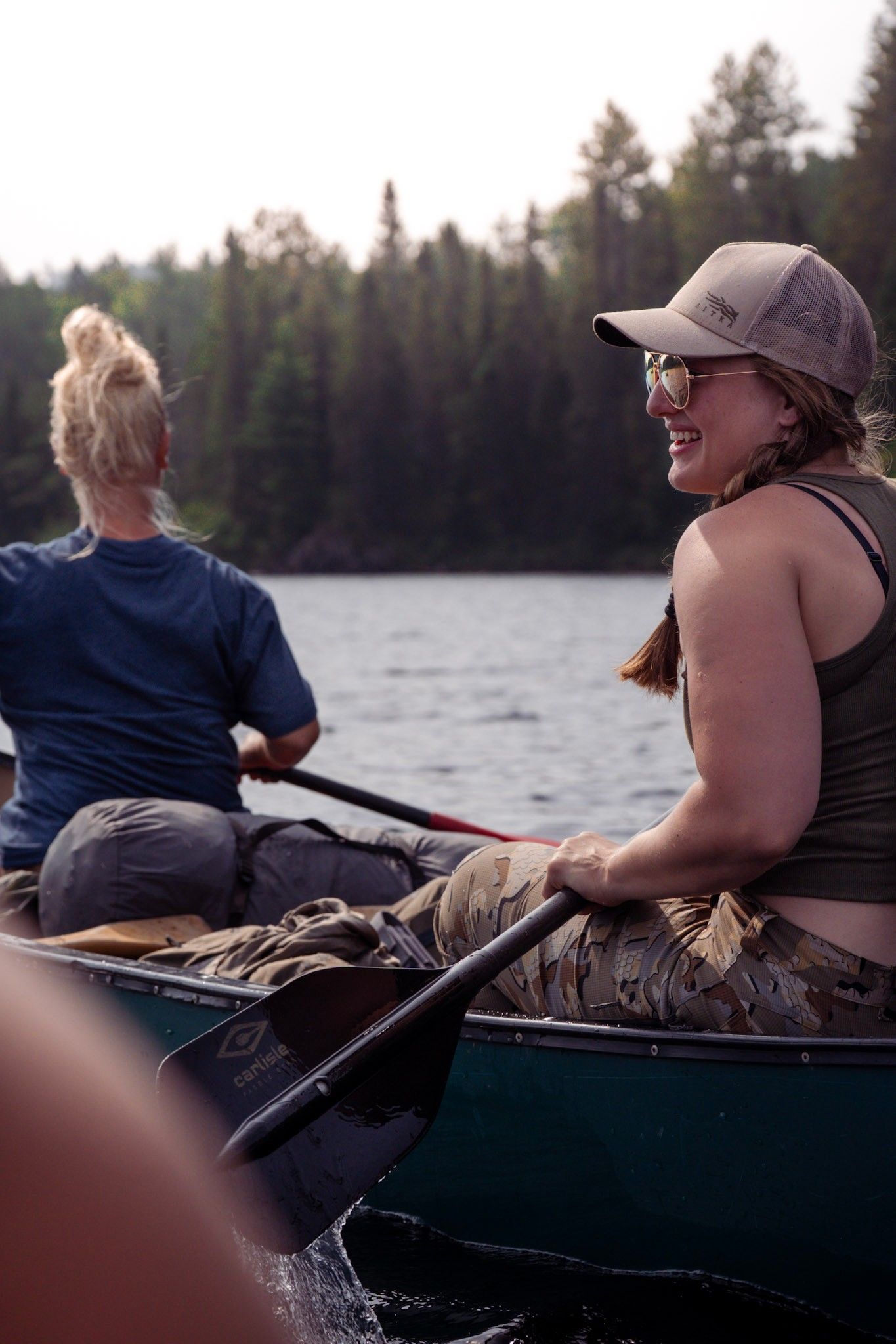 Two people paddling a canoe on a lake at dusk with evergreen trees lining the forested shoreline in the background.