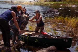 Group of people working together to lift a canoe near a rocky riverbank with tall grass.