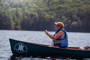 A paddler in an orange cap and blue life vest checks their phone while sitting in a green canoe on a calm lake.