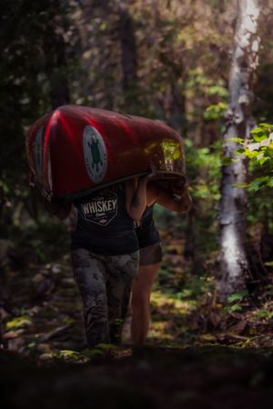 Someone wearing a whiskey t-shirt carries a large red canoe through a dark wooded forest trail.