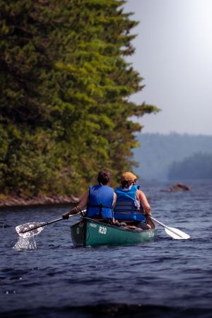 Two people paddling a green canoe on a scenic lake with evergreen trees along the shoreline in summer.
