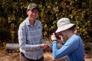 Two hikers wearing hats and long sleeves interact outdoors against a backdrop of autumn foliage.