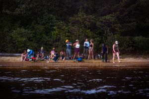 A group of people gather along a shoreline at dusk, silhouetted against dark water and forested background.