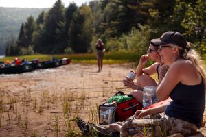 People relax on the beach with kayaks and gear while enjoying a sunny day near a forested lakeshore.