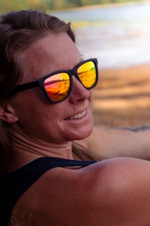 A person wearing reflective orange sunglasses smiles while relaxing outdoors during golden hour.