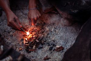 Hands tending to a small glowing campfire with embers and ashes in the dark.