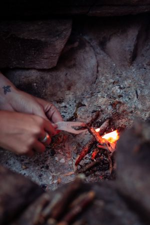 Hands tending to a small campfire with glowing embers and flames amongst rocks and ashes.