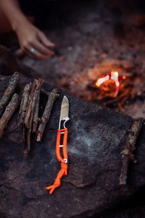 A camping knife with orange handle rests on a log near glowing embers and sticks in a dark outdoor setting.
